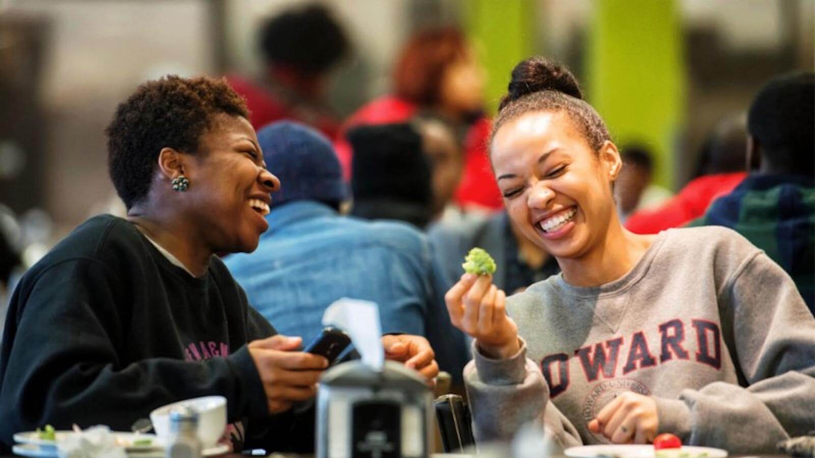 Two women in Howard sweatshirts sitting at a lunch table, laughing.