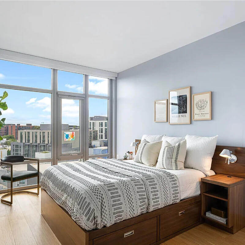 Furnished apartment at The Lanes, showing a bed, nightstand, and chair by a beautiful window with a view of the city.