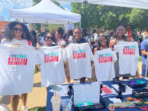 5 female students standing at the IVPP resources table, holding "HU Stands Against Sexual Assault" Shirts and smiling.